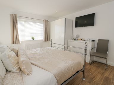 A bedroom with a bed a window with curtains a wardrobe a dresser with items on top a chair and a wall-mounted television at The Gables in Tywyn