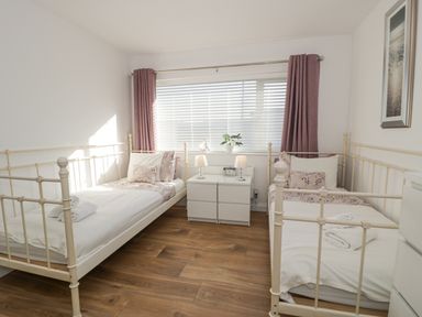 A bedroom with two single metal frame beds a white nightstand with lamps and a clock between them and a window with blinds and purple curtains at The Gables in Tywyn