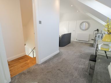 An upper hallway with gray carpet a staircase with wooden steps a gray sofa a wall-mounted radiator a round mirror and a mirrored console table with a yellow vase with flowers at The Gables in Tywyn