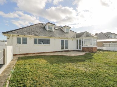 A backyard with grass and a paved patio leading to a white house with dormer windows and a sunroom at The Gables in Tywyn