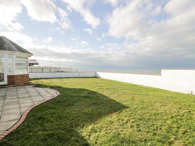 A fenced grassy yard with a paved patio next to a building near the coast at The Gables in Tywyn