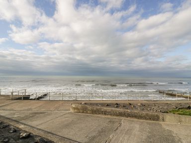 A concrete pathway with a metal railing overlooking sea waves under a cloudy sky at The Gables in Tywyn