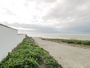 A concrete pathway leading to the sea with green plants on both sides and a white wall on the left at The Gables in Tywyn