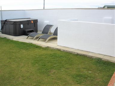 An outdoor area with two lounge chairs and a covered hot tub on a paved surface next to a grass lawn at The Gables in Tywyn