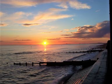 A sunset over the ocean with wooden groynes along the shore and a paved walkway at The Gables in Tywyn