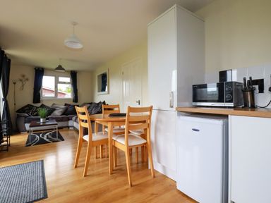 A kitchen and living area with a wooden dining table and chairs a microwave on countertop a gray sofa and window with curtains at Gwanda in Tregolds near St Merryn