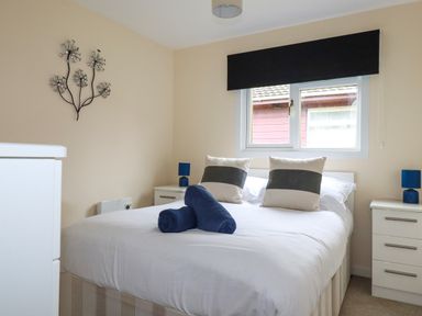 A bedroom with a double bed white bedding two pillows blue rolled towels two bedside tables with blue lamps and a window with a black blind at Gwanda in Tregolds near St Merryn