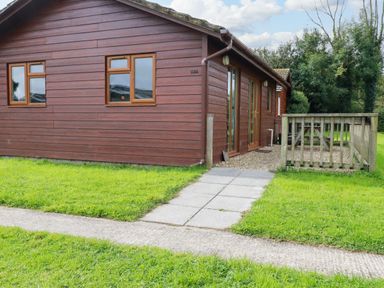 An exterior view of a wooden cabin with windows and a door leading to a small fenced patio with grass surrounding it at Gwanda Tregolds near St Merryn