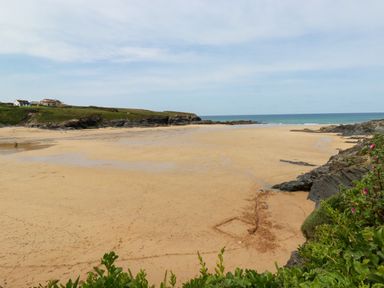 A sandy beach with a few rocky outcrops green vegetation in the foreground and houses on a grassy hill in the background at Gwanda Tregolds near St Merryn