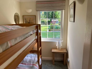 A bedroom with wooden bunk beds a small wooden side table with a lamp a window overlooking a garden and framed pictures on the wall at October House Holiday Cabin in Damerham