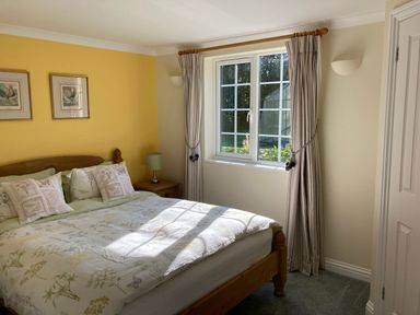 A bedroom with a wooden bed with floral bedding a bedside table with a lamp and a window with curtains at October House Holiday Cabin in Damerham