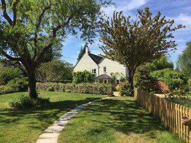 A garden with trees a stone pathway and a white house behind a wooden fence at October House Holiday Cabin in Damerham