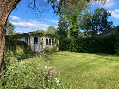 A garden with a green lawn flowers a tree and a small white building covered with plants at October House Holiday Cabin in Damerham