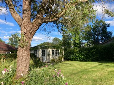 A garden with a large tree flowers and a small building with windows and a door at October House Holiday Cabin in Damerham
