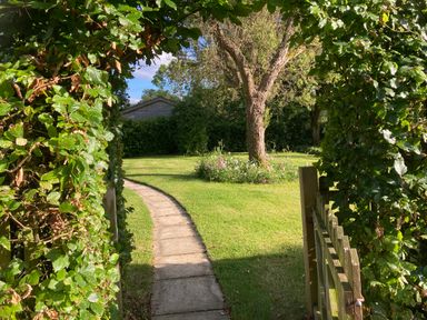 A stone pathway leading through a wooden gate and green hedge to a yard with a tree and flower bed at October House Holiday Cabin in Damerham
