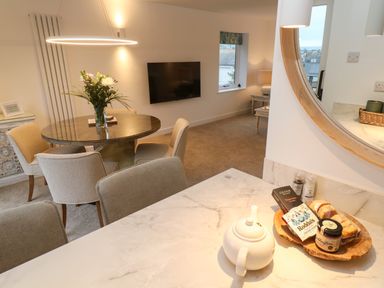 A dining area with a round table and chairs a bouquet of flowers on the table a wall-mounted TV and a countertop with a teapot and snacks at Fal View in Falmouth