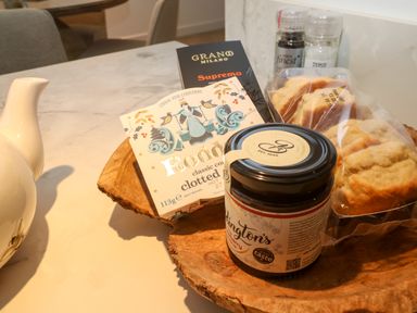 A wooden platter with a jar of jam, a box of clotted cream, a chocolate bar, a plastic bag of scones, and salt and pepper shakers on a marble countertop at Fal View in Falmouth