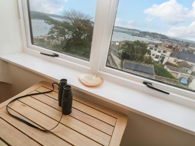 A wooden table with binoculars by a window overlooking houses trees and water at Fal View in Falmouth