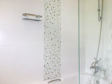 A bathroom with a bathtub that has a patterned glass shower screen a handheld shower head and tiled white walls with a mosaic strip at Trestar in Polzeath