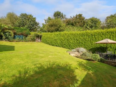 A garden with a large lawn hedge trees outdoor seating with umbrella and a small shed at Trestar in Polzeath