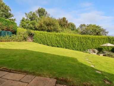 A backyard with a grassy lawn a small shed in the corner a hedge along the back and a patio umbrella at Trestar in Polzeath