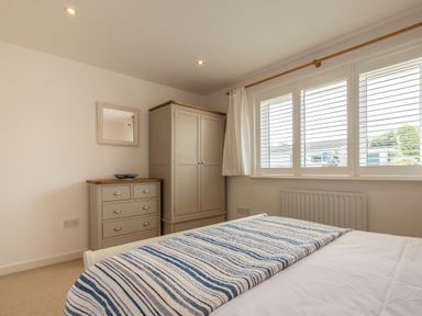 A bedroom with a wardrobe, chest of drawers, mirror, window with blinds and a bed with a striped blanket at Trestar in Polzeath