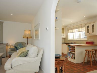 A living room with a white sofa and lamps next to an open plan kitchen with wooden stools and shelves at Trestar in Polzeath