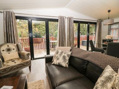 A living room with leather sofa patterned armchair glass dining table and large sliding doors overlooking a wooden deck at Somerford Lodge Langdale 7 White Cross Bay near Windermere