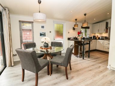A dining area with a glass table and four chairs next to a kitchen with a bar counter and stools at Somerford Lodge Langdale 7 White Cross Bay near Windermere