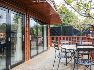 An outdoor wooden deck with a round table and four metal chairs near sliding glass doors at Somerford Lodge Langdale 7 White Cross Bay near Windermere