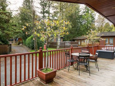 An outdoor wooden deck with potted plants and a table with chairs overlooking a path and wooden cabins at Somerford Lodge Langdale 7 White Cross Bay near Windermere