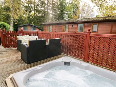 An outdoor deck with a hot tub and black wicker seating near wooden cabins surrounded by trees at Somerford Lodge Langdale 7 White Cross Bay near Windermere