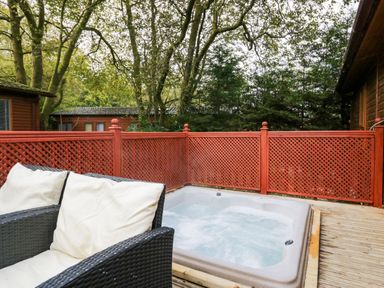 An outdoor deck with two cushioned chairs and a hot tub surrounded by a red wooden fence at Somerford Lodge Langdale 7 White Cross Bay near Windermere