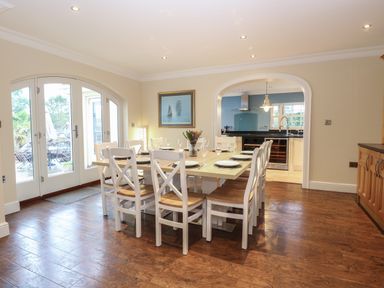 A dining area with a wooden table and white chairs set with plates and glasses adjacent to a kitchen at The Little House in Horning