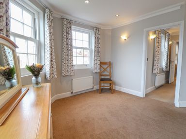 A room with beige carpet a wooden chair a wooden dresser with a mirror and two windows with floral curtains at The Little House in Horning