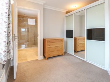 A bedroom with a wooden chest of drawers a sliding door wardrobe with mirrors and a doorway leading to a tiled bathroom at The Little House in Horning
