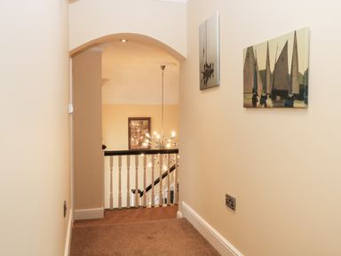 A hallway with beige walls and carpet leading to a staircase with white railing and pictures on the wall at The Little House in Horning