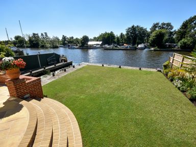 A garden with grass lawn and flower pots overlooking a river with boats at The Little House in Horning