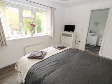 A bedroom with a bed covered with a dark grey blanket and towels on top a chest of drawers with a television and a window with curtains and flowers in a vase at Butterfly Lodge in Tunstall near Catterick North Yorkshire
