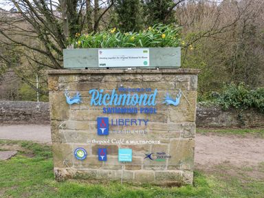 A stone sign with blue lettering welcoming to Richmond Swimming Pool with logos and a planter with flowers on top at Butterfly Lodge in Tunstall near Catterick North Yorkshire