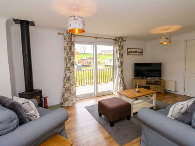 A living room with gray sofas a brown ottoman coffee table wood burner and sliding doors overlooking a lawn at Butterfly Lodge in Tunstall near Catterick North Yorkshire