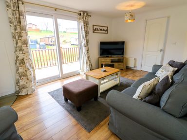 A living room with a gray sofa and cushions a brown ottoman a wooden coffee table a flat screen tv on a wooden stand and sliding glass doors with patterned curtains at Butterfly Lodge in Tunstall near Catterick North Yorkshire