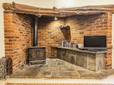 A brick fireplace area with a wood burning stove a television a stone bench with books and a log beam at The Log Cabin in Sheldon Blackdown Hills