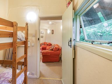 A bedroom with wooden bunk beds connected to a living room with orange armchairs and a window showing greenery at The Log Cabin in Sheldon Blackdown Hills