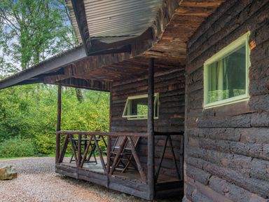 A wooden cabin porch with two chairs and wooden railing surrounded by greenery at The Log Cabin in Sheldon Blackdown Hills