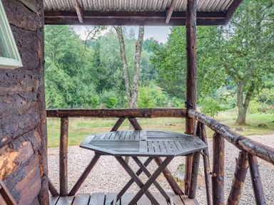 A wooden porch with a round table overlooking trees and grass at The Log Cabin in Sheldon Blackdown Hills