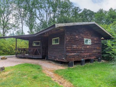 A wooden log cabin with small windows and a covered porch with chairs surrounded by trees and grass at The Log Cabin in Sheldon Blackdown Hills