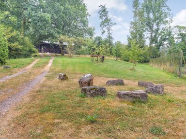 A gravel driveway leading to a cabin surrounded by trees and grassy area with a circle of large stones at The Log Cabin in Sheldon Blackdown Hills