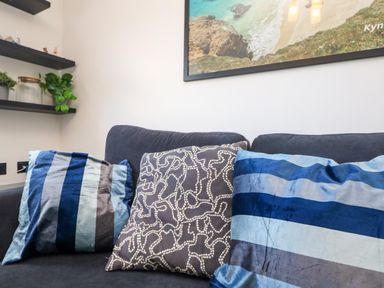 A black sofa with three patterned cushions and shelves with plants and jars on a white wall at Savoy View in Mullion