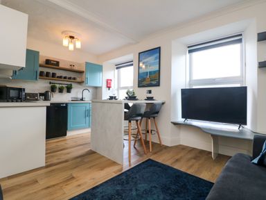 A kitchen and dining area with blue cabinets stools and a TV near windows at Savoy View in Mullion